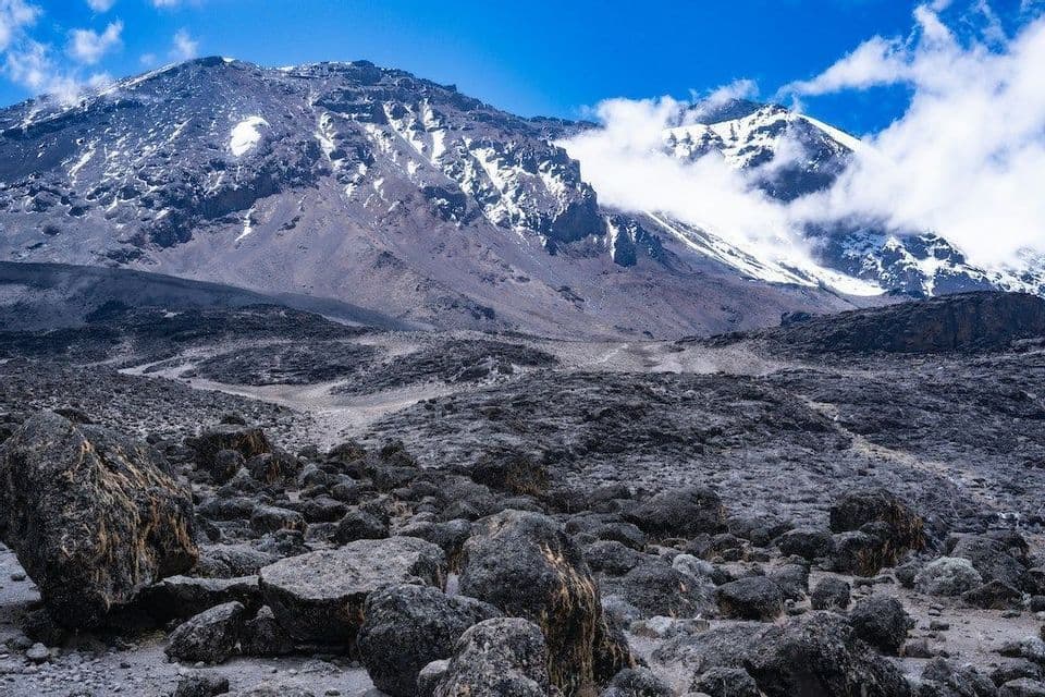Un vaste paysage rocheux s'étend vers des montagnes enneigées imposantes sous un ciel bleu éclatant, partiellement nuageux.