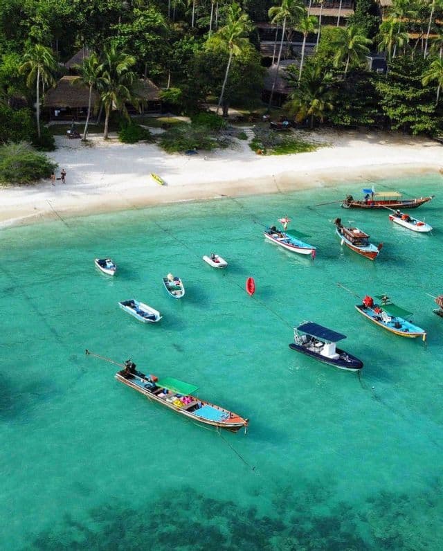 Una vista aérea muestra muchos barcos anclados en aguas turquesas poco profundas junto a una playa de arena blanca con palmeras.