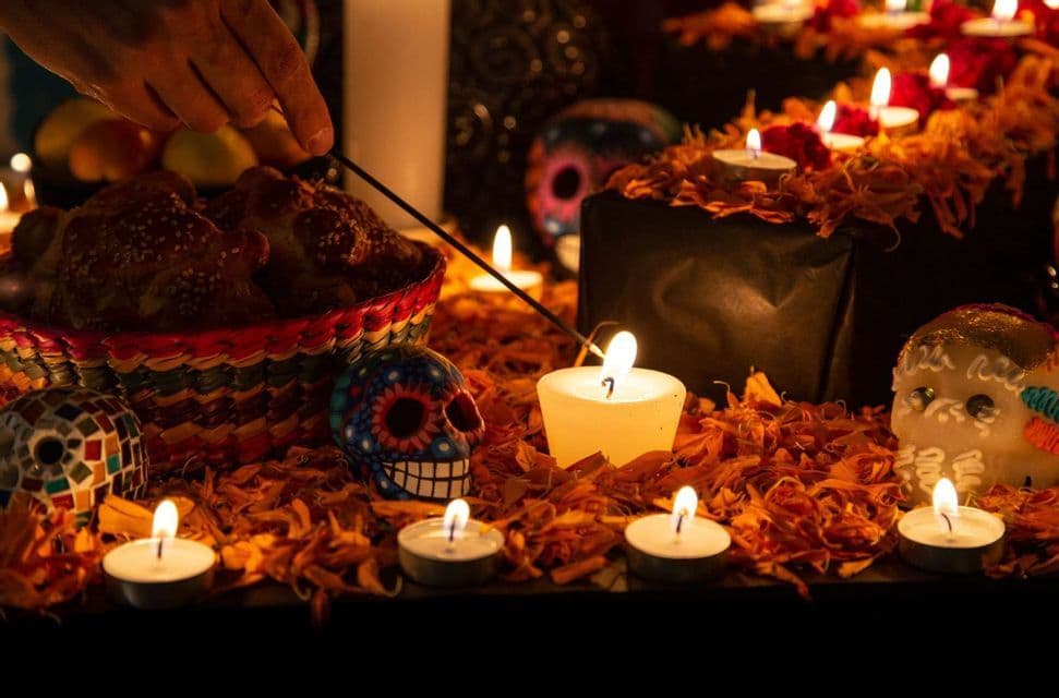A hand lights an incense stick over a Dia de los Muertos altar with candles, sugar skulls, and marigold petals.