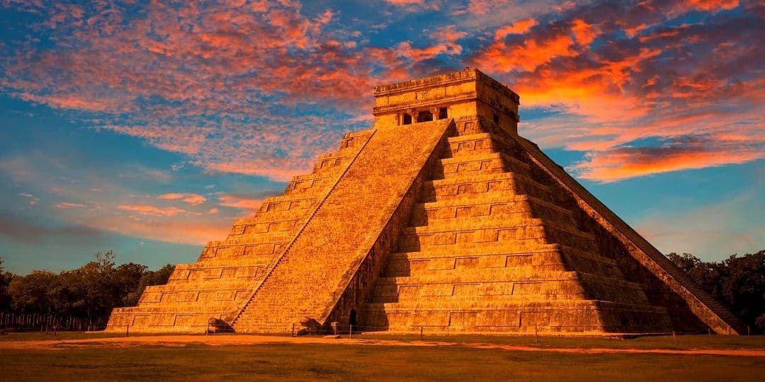 Una piramide a gradoni di pietra è illuminata dalla luce dorata del sole, stagliandosi su un drammatico cielo al tramonto con nuvole arancioni e blu.