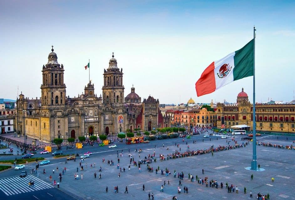 A large Mexican flag waves over a historic city square bustling with people, with an ornate cathedral in the background.