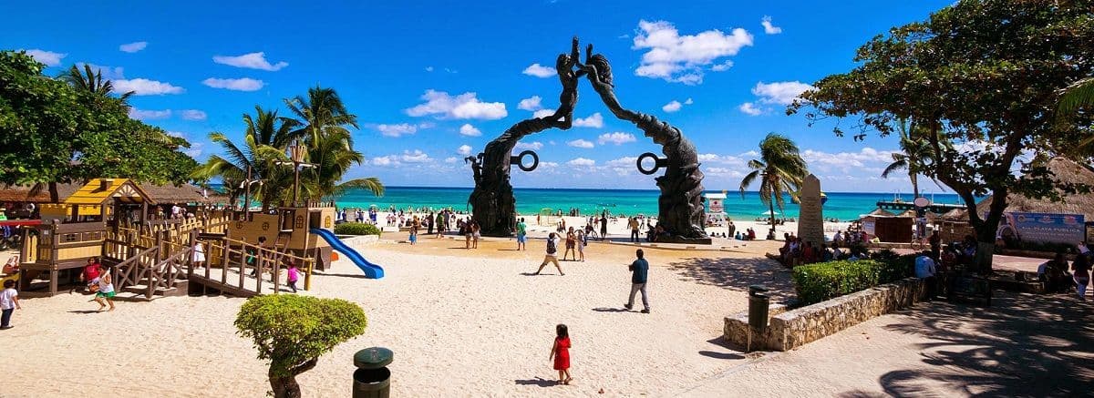Una grande scultura in bronzo si inarca sopra una trafficata spiaggia di sabbia bianca, accanto a un parco giochi, con acqua turchese sotto un cielo azzurro.