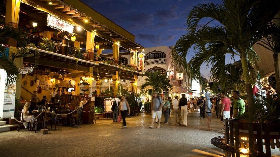 A bustling street scene at dusk, with people walking along a paved road lined with illuminated restaurants and palm trees.