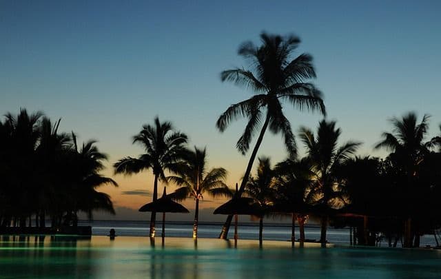 Silhouettierte Palmen und Strohschirme stehen bei Sonnenuntergang an einem Infinity-Pool mit dem Meer im Hintergrund.