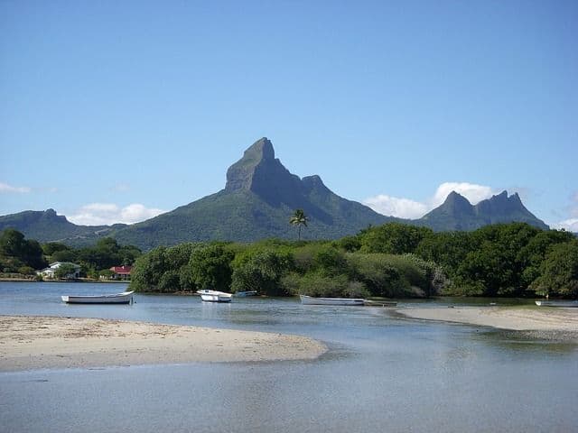 Blick auf eine ruhige Bucht mit mehreren Booten, die nahe eines Sandstrandes ankern, vor dem Hintergrund üppiger, zerklüfteter Berge unter klarem blauem Himmel.
