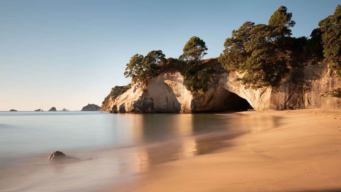 A large cave opening in a rocky cliff on a sandy beach, with calm water reflecting the clear sky at sunrise.