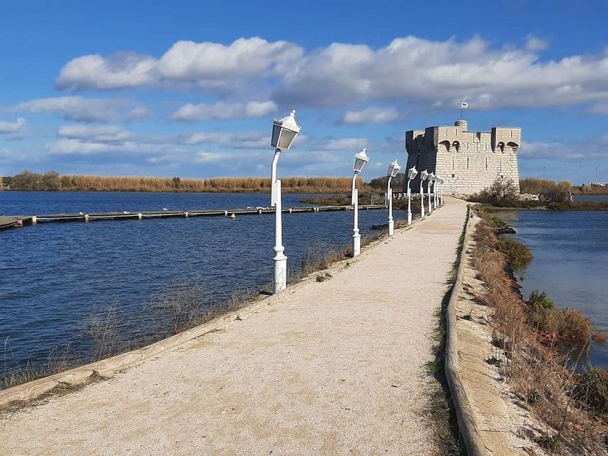 Une chaussée sablonneuse bordée de lampadaires blancs mène à un fort en pierre entouré d'eau, sous un ciel bleu parsemé de nuages.