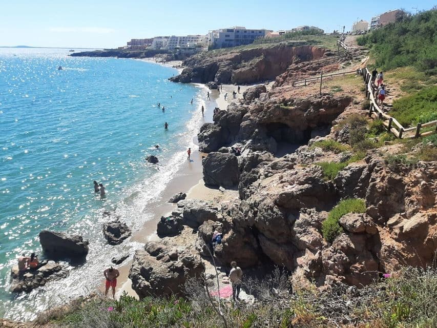 Vue plongeante sur une plage ensoleillée, avec des gens nageant dans la mer bleue et se relaxant le long du littoral rocheux.