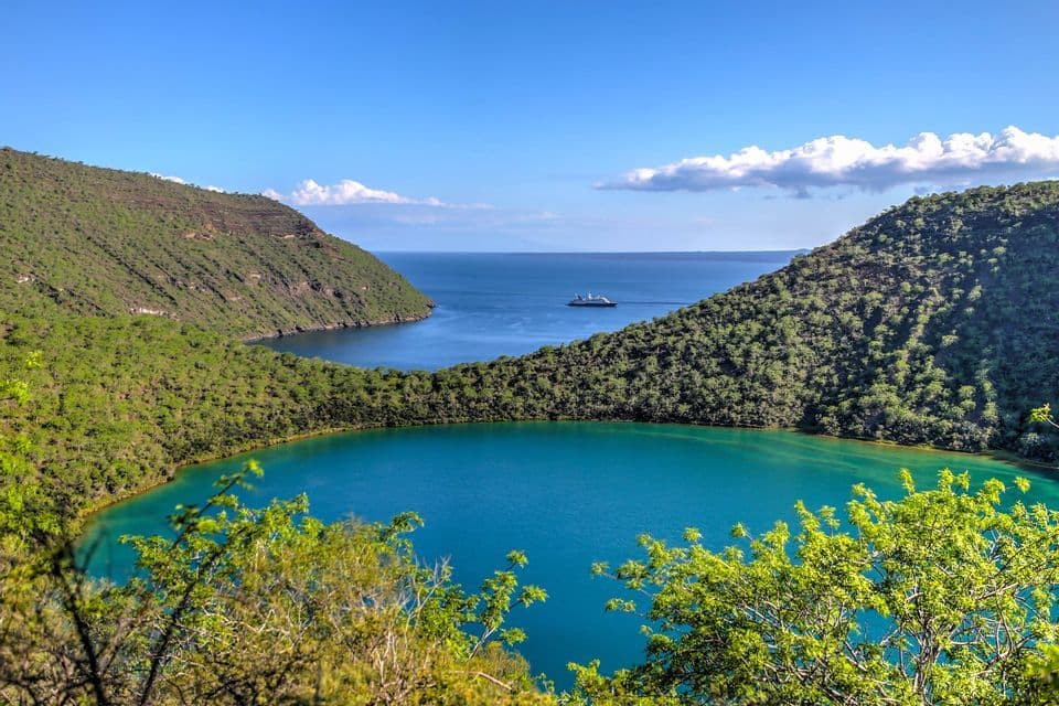 Un lago de cráter turquesa se anida entre colinas verdes, con el océano y un crucero visibles a la distancia bajo un cielo azul.