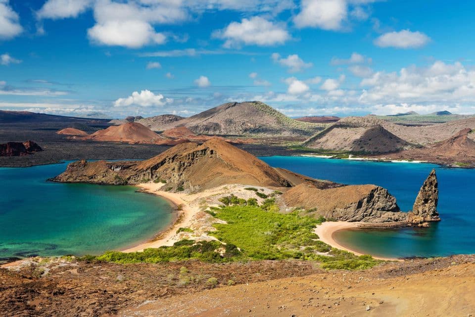 Vista aérea de islas volcánicas con agua turquesa en una bahía, un prominente peñón marino y playas de arena bajo un cielo azul nublado.