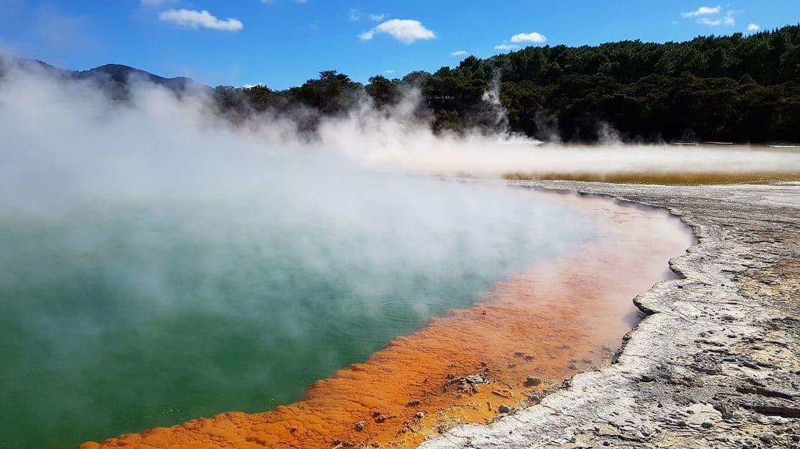 De la vapeur s'élève d'une piscine géothermique aux eaux vertes et oranges vives, bordée par une rive rocheuse avec une forêt en arrière-plan.
