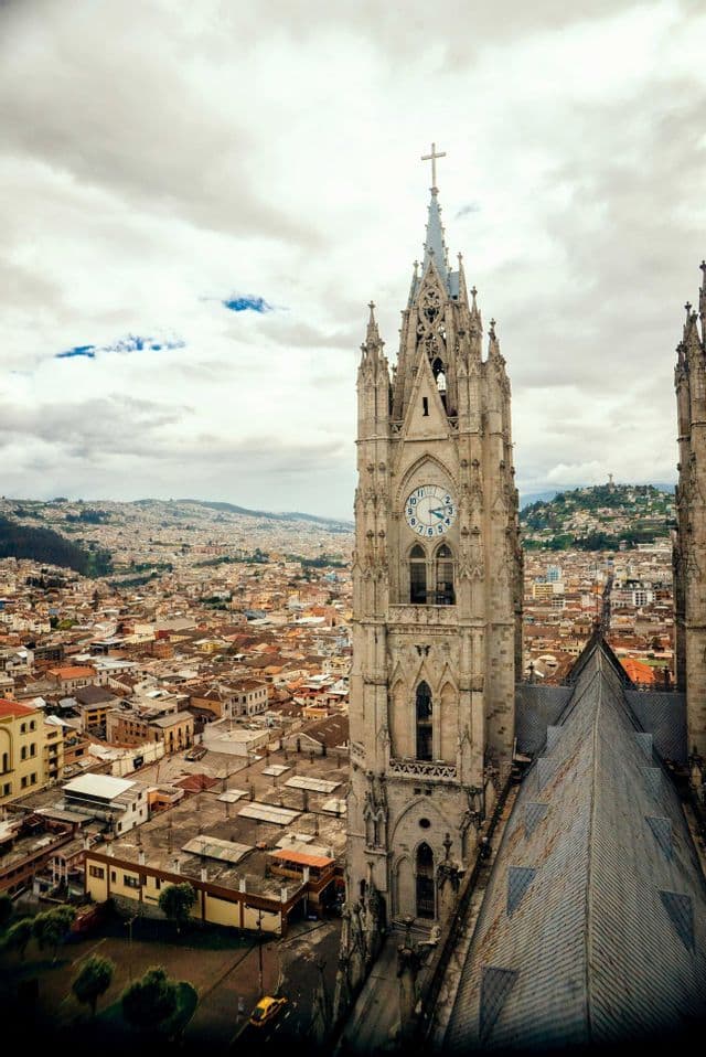 Un campanario gótico ornamentado de una catedral domina una extensa ciudad con colinas distantes bajo un cielo nublado.
