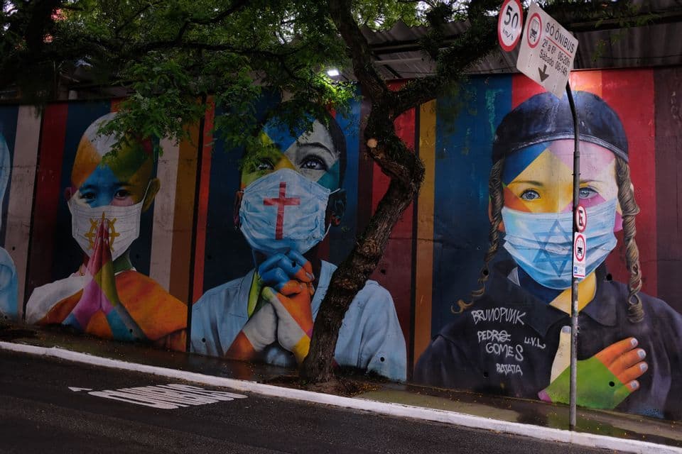 Un murale colorato di street art su un muro cittadino che mostra tre bambini che indossano mascherine con diversi simboli religiosi.