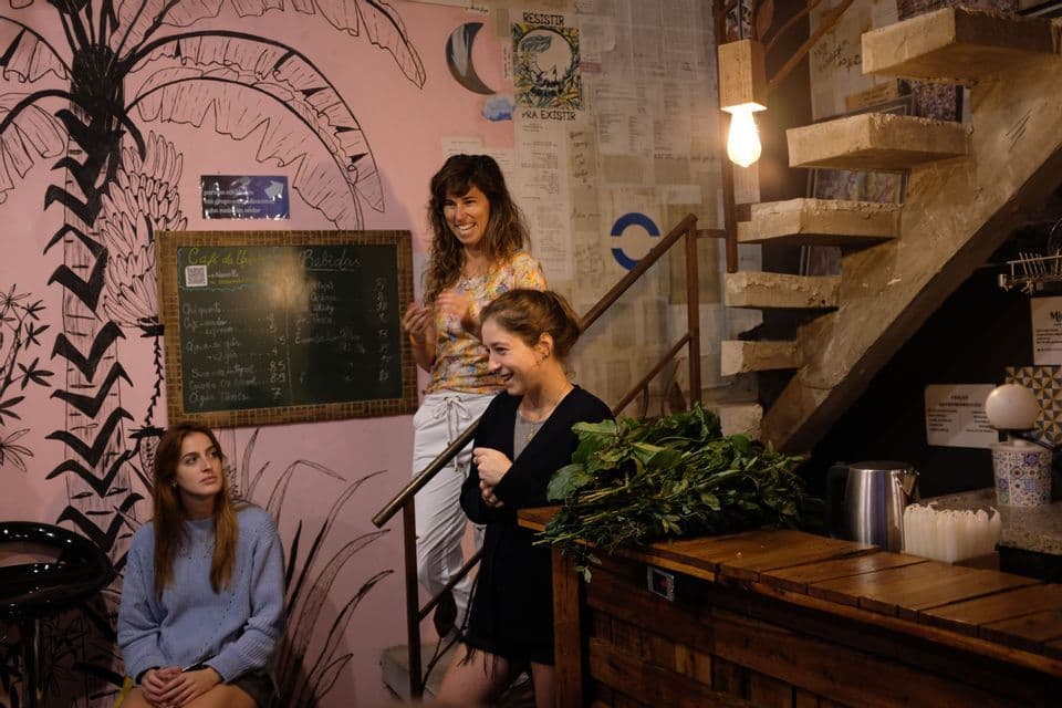 Trois femmes d'un groupe WeRoad rient dans un café avec une fresque murale rose, l'une tenant un grand bouquet d'herbes.