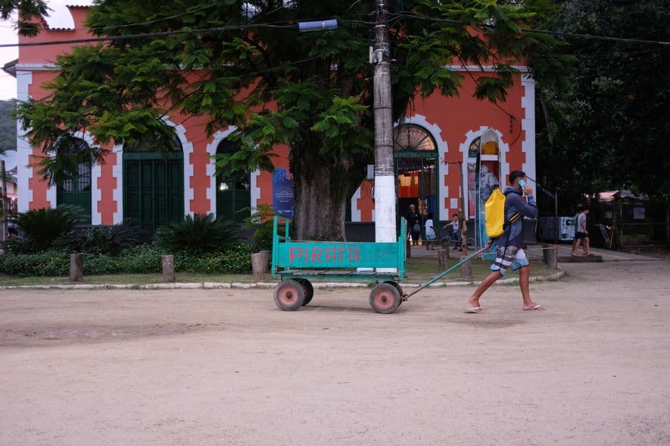 Un hombre con una mochila amarilla arrastra un carrito verde por un camino de arena frente a un edificio colonial naranja.