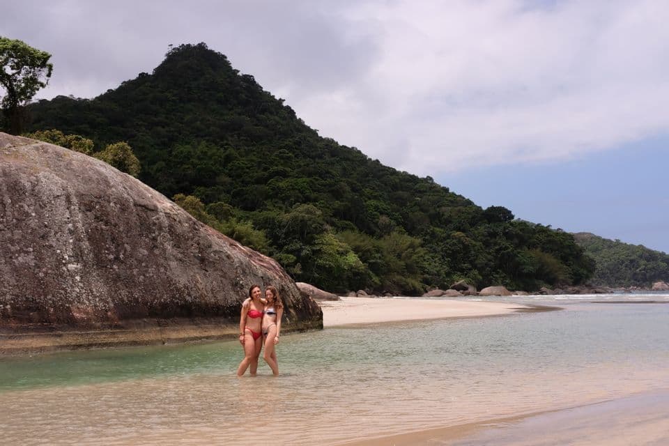 Dos mujeres en bikini de un viaje en grupo de WeRoad posan sonriendo en las aguas poco profundas de una playa, con una gran roca y una colina boscosa detrás de ellas.