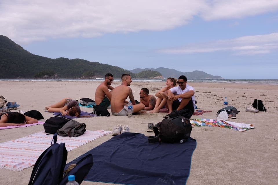 Un grupo de WeRoad relajándose en toallas en una amplia playa de arena, con verdes colinas arboladas al fondo, al otro lado del agua.