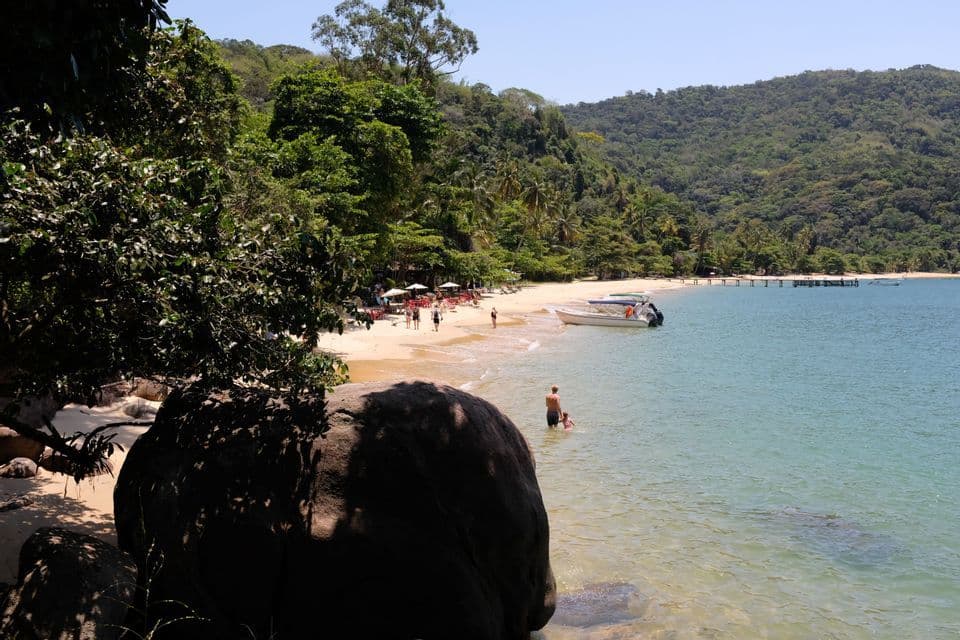 Gente relajándose en una playa de arena junto a aguas turquesas, con un barco amarrado y una exuberante colina cubierta de árboles al fondo.