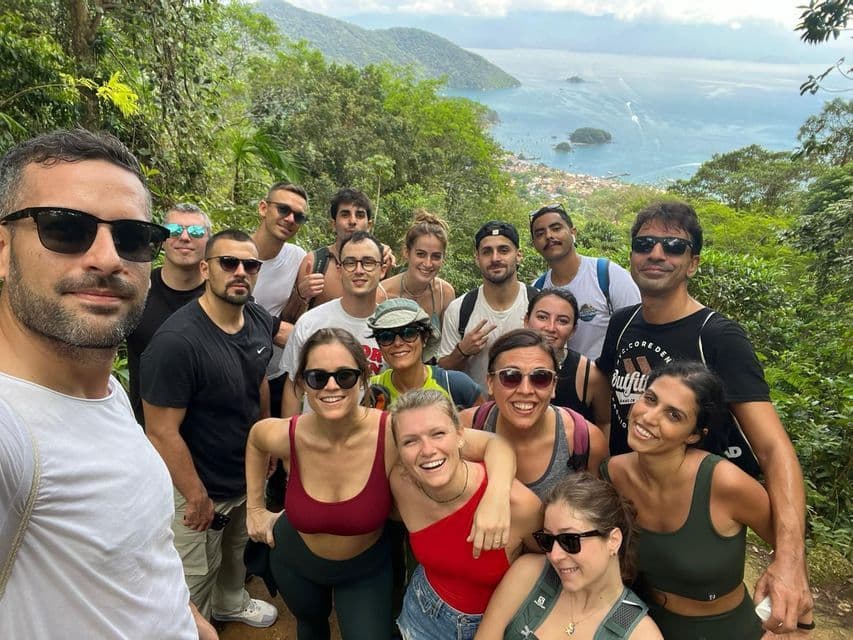 Un gruppo WeRoad in viaggio posa per un selfie su una rigogliosa collina verde con vista su una baia costiera e le montagne.