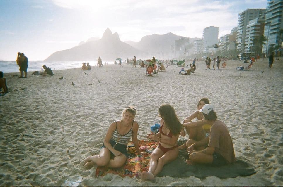 Un groupe WeRoad de quatre personnes assises sur des serviettes sur une plage bondée, avec des montagnes et une ville en arrière-plan.