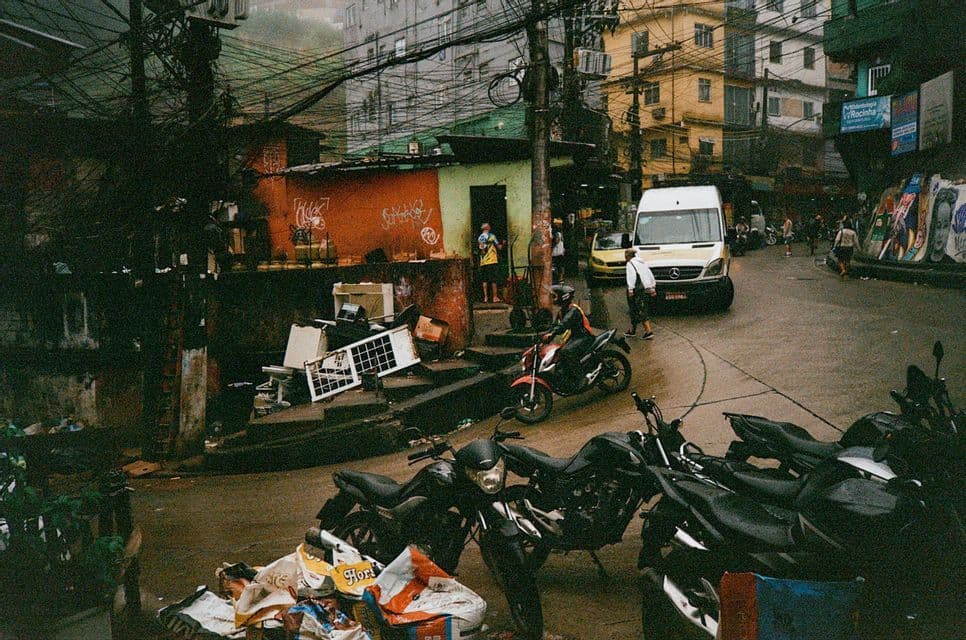 Motociclette parcheggiate su una strada affollata e bagnata con un furgone, edifici coperti di graffiti e numerosi cavi aerei aggrovigliati.