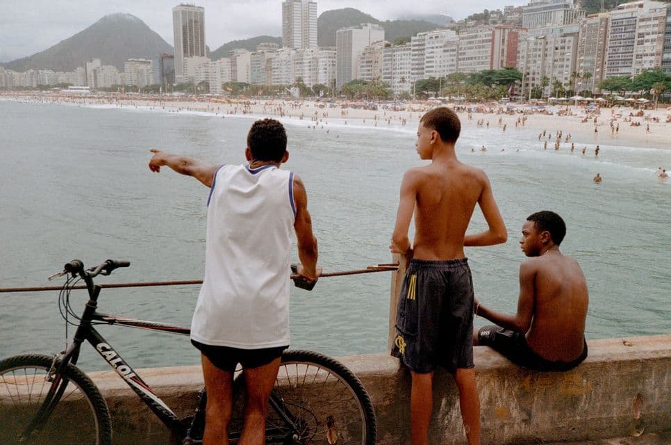 Tre giovani visti di spalle con una bicicletta su un molo, che osservano una spiaggia affollata e lo skyline di una città.