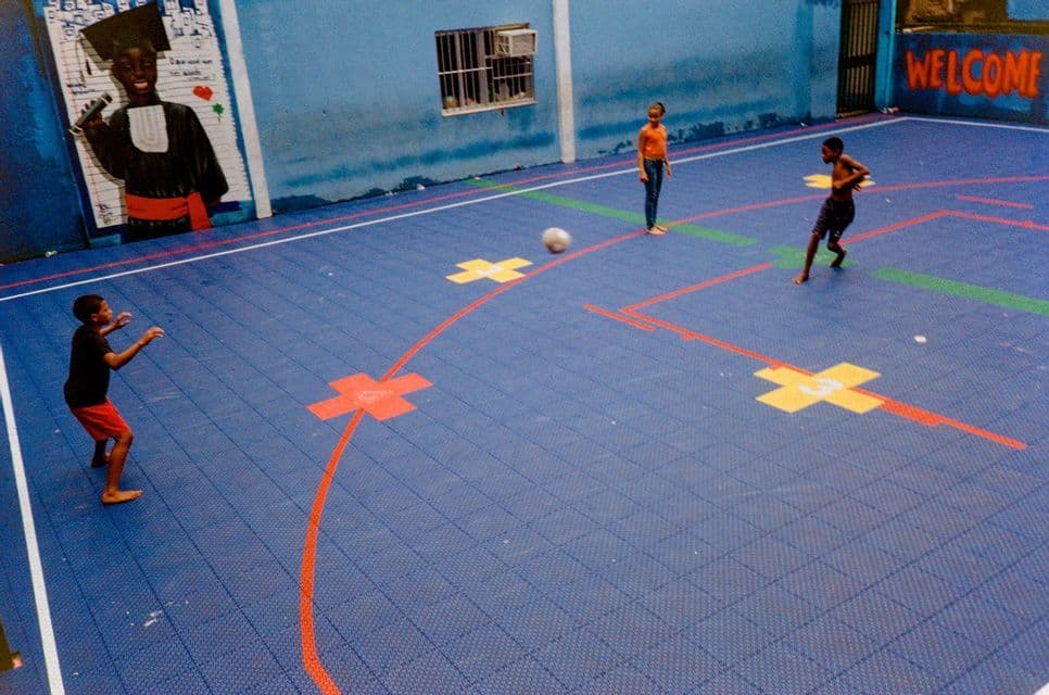 Drei barfüßige Kinder spielen mit einem Ball auf einem blau gefliesten Indoor-Sportplatz mit einem großen Wandgemälde.