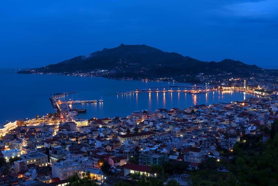 Una vista dall'alto di una città costiera illuminata al crepuscolo, con le sue luci che si riflettono in una baia tranquilla e una montagna sullo sfondo.