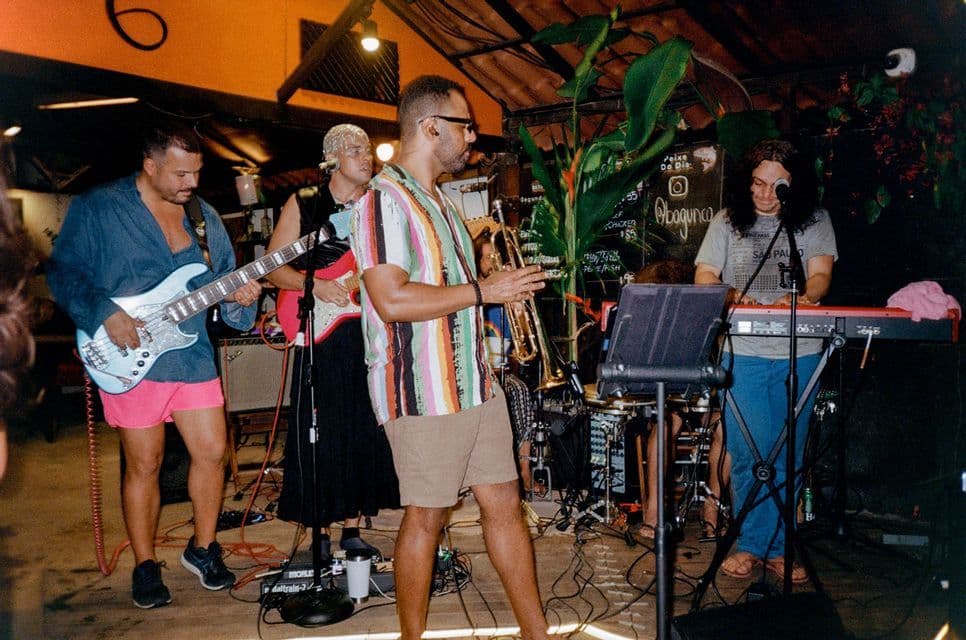 Un groupe de quatre musiciens, avec un bassiste, un guitariste, un trompettiste et un claviériste, se produit sur une scène faiblement éclairée.