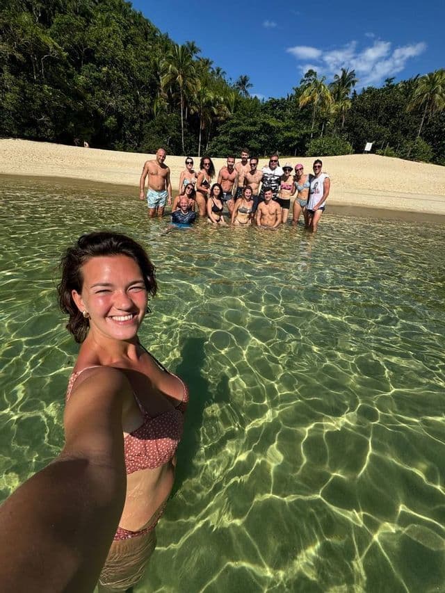Una mujer en bikini se toma una selfie con su grupo de viaje de WeRoad en las aguas claras y poco profundas de una playa tropical.
