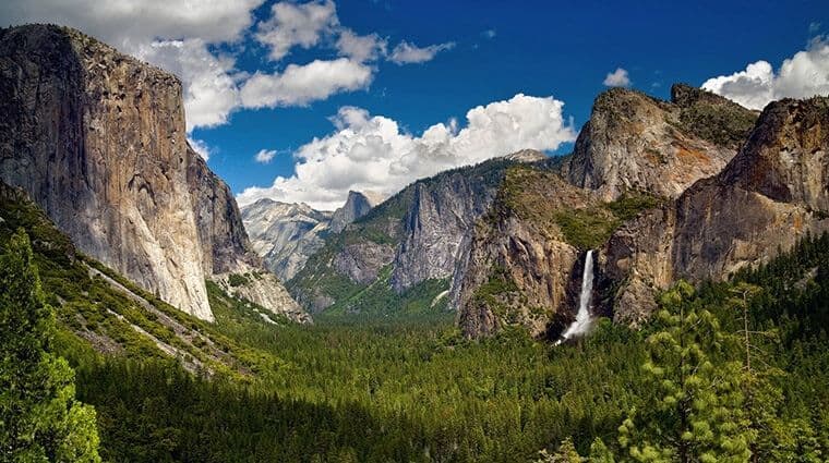 Un'ampia valle con una fitta foresta verde, fiancheggiata da imponenti scogliere di granito e una cascata, sotto un cielo azzurro con nuvole.