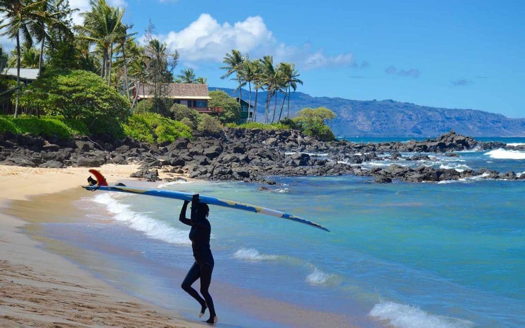 Un surfista con muta nera porta una longboard lungo una spiaggia tropicale con acqua turchese.