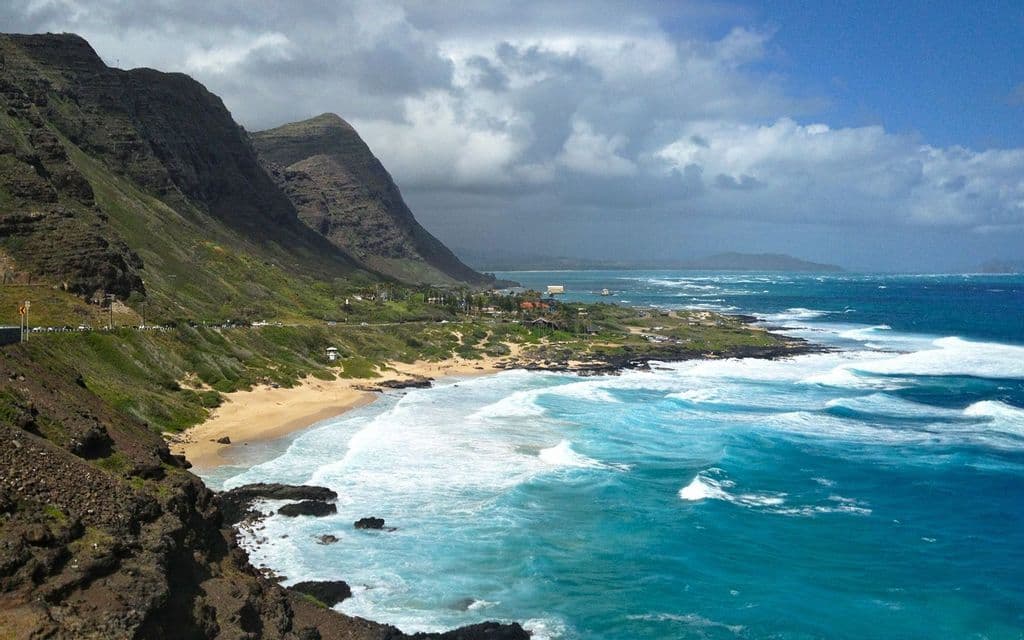 Una vista dall'alto di una costa aspra e verde dove le onde turchesi dell'oceano si infrangono su una spiaggia sabbiosa sotto un cielo parzialmente nuvoloso.