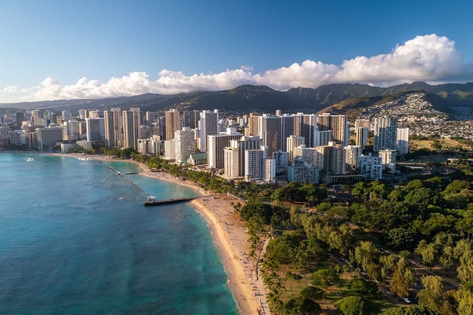 Veduta aerea di uno skyline cittadino accanto a una spiaggia sabbiosa e un mare turchese, con montagne verdi sotto un cielo blu.