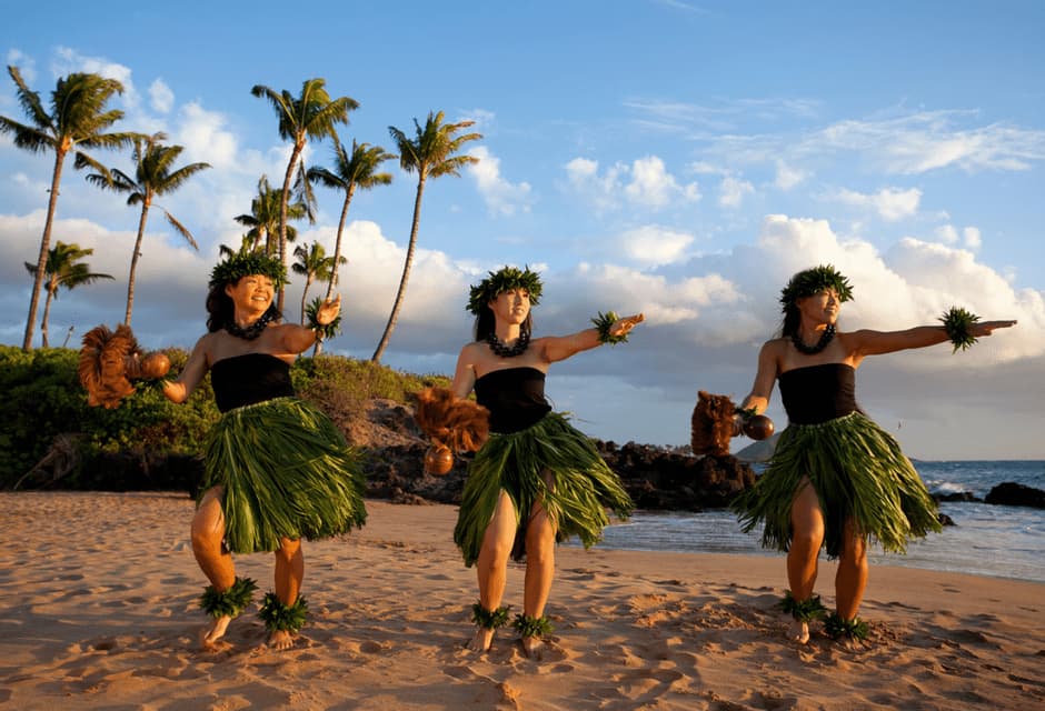 Tre donne in gonne di foglie tradizionali e ghirlande eseguono una danza hula su una spiaggia sabbiosa con palme sullo sfondo.