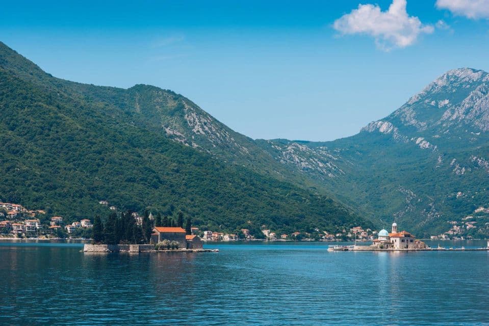 Due piccole isole con edifici sorgono su uno specchio d'acqua calmo, con montagne verdi e boscose che si ergono sullo sfondo sotto un cielo azzurro.