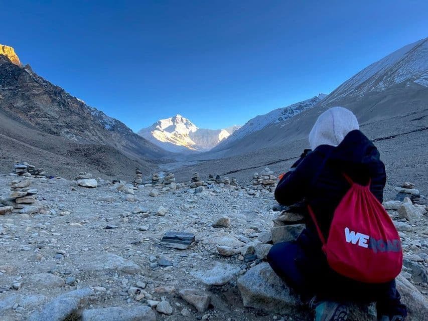 Una persona con uno zaino rosso WeRoad è inginocchiata su un terreno roccioso, ammirando una catena montuosa innevata e illuminata dal sole in lontananza.