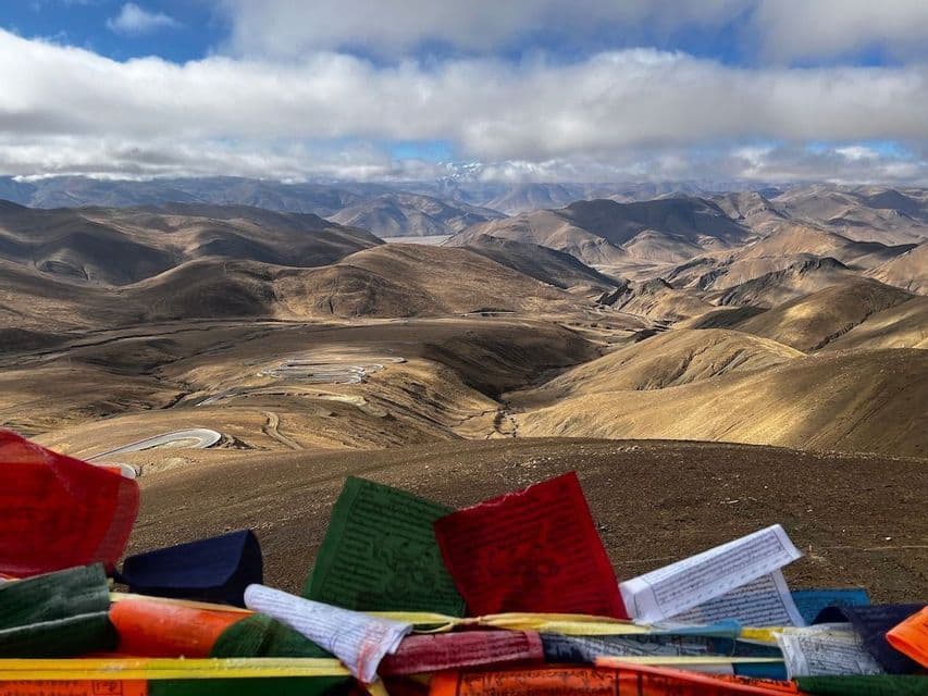 Bandiere di preghiera colorate si affacciano su una strada tortuosa attraverso un vasto paesaggio di montagne aride e ondulate sotto un cielo nuvoloso.