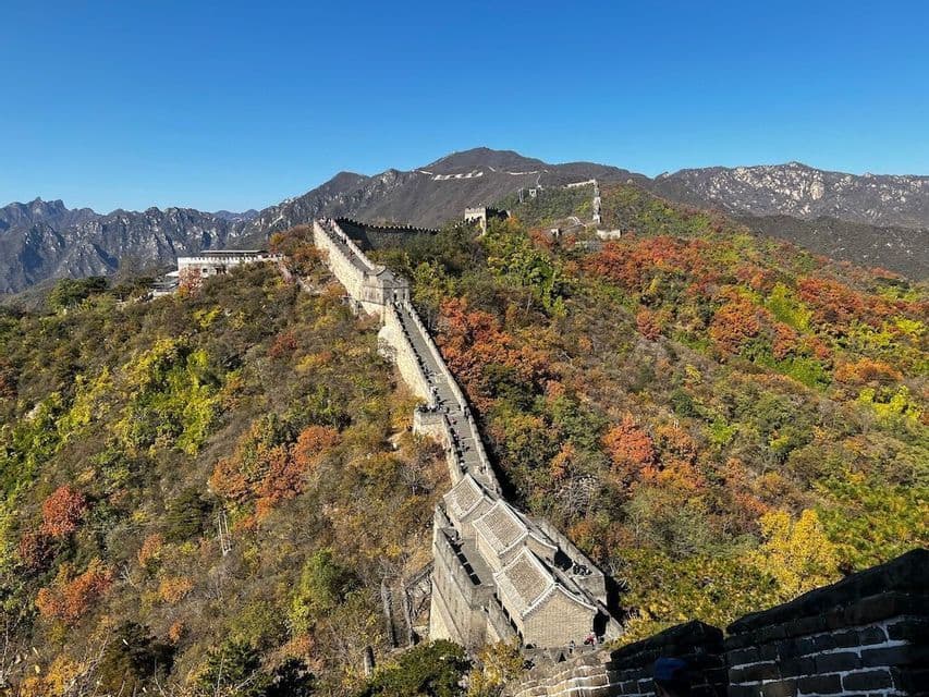 Una grande muraglia di pietra si snoda tra dolci montagne ricoperte di foglie autunnali colorate, con persone che la percorrono sotto un cielo sereno.