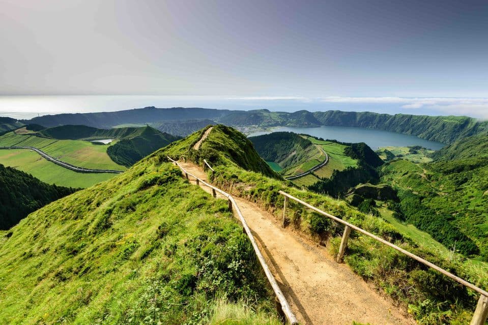 Un sentier de randonnée en terre battue avec une rambarde en bois longe une crête herbeuse surplombant une vallée avec un lac de cratère et l'océan.