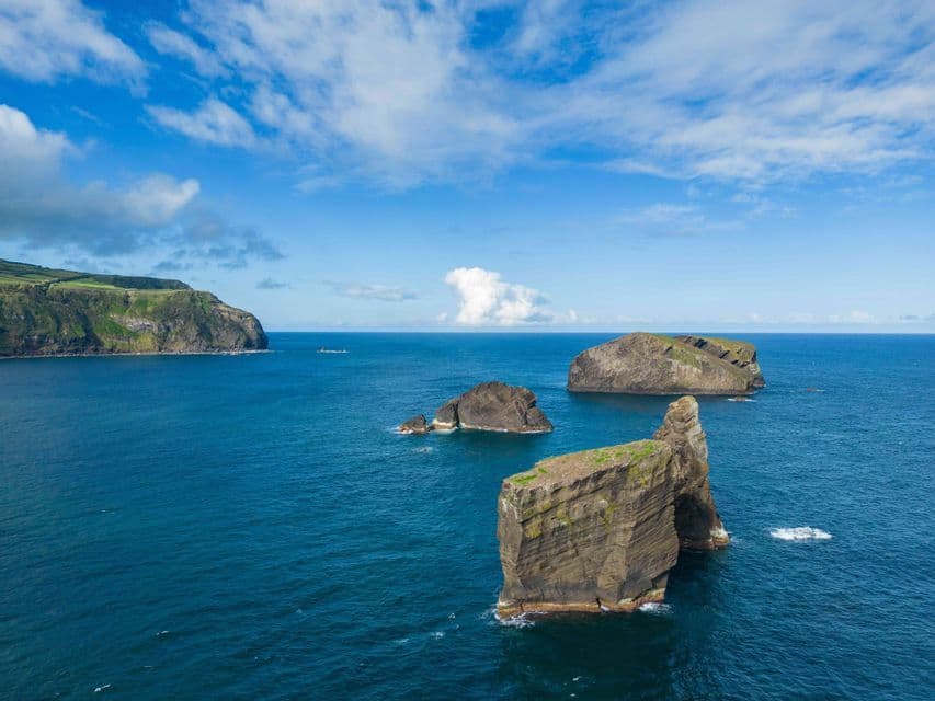 Vue aérienne de grands rochers marins, dont un avec une arche naturelle, dans l'océan bleu, le long d'une côte verte et escarpée.