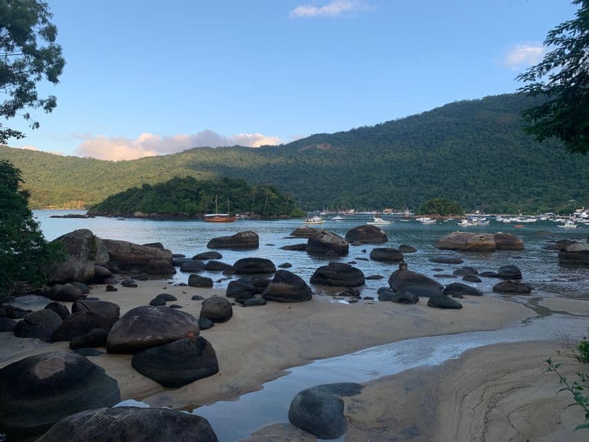 Grandes rocas oscuras en una playa de arena junto a una bahía tranquila repleta de barcos, con una gran colina verde al fondo.