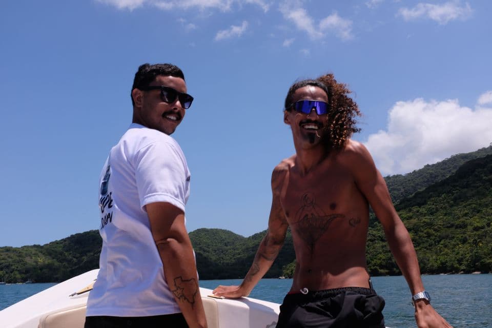 Dos hombres de un viaje en grupo de WeRoad sonriendo en un barco, con una costa verde y cielo azul de fondo.