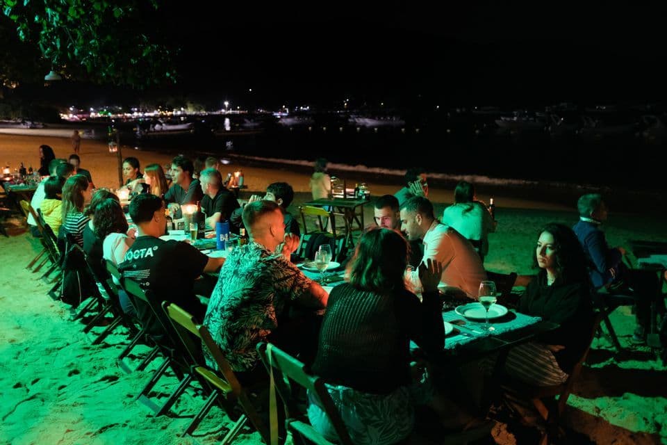 Un viaje en grupo de WeRoad cenando en una mesa larga en una playa de arena por la noche, con el mar y barcos al fondo.