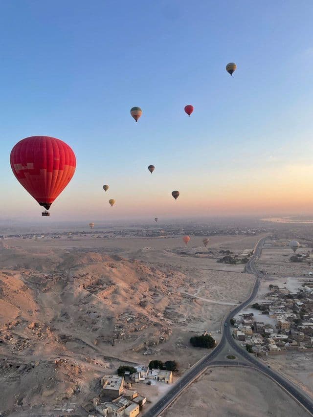Varios globos aerostáticos vuelan sobre un vasto paisaje desértico con un pequeño pueblo al amanecer.