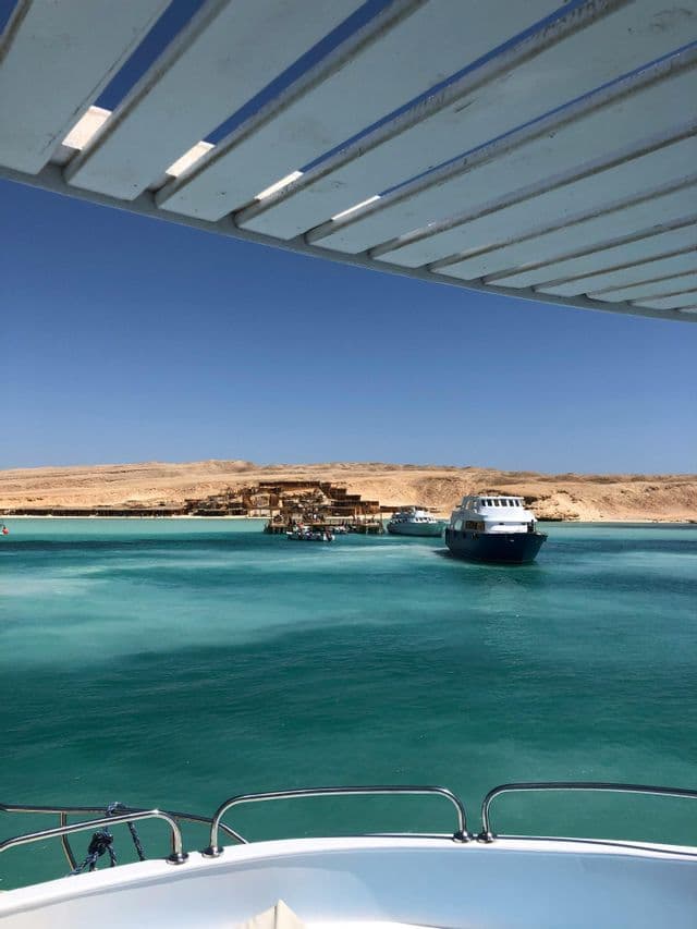 Vista desde la cubierta de un barco de varias embarcaciones amarradas en agua turquesa junto a una costa arenosa y desértica.
