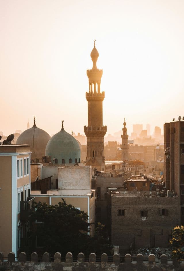 Cúpulas de mezquita y un alto minarete se alzan sobre un denso paisaje urbano bañado por la luz dorada del atardecer.