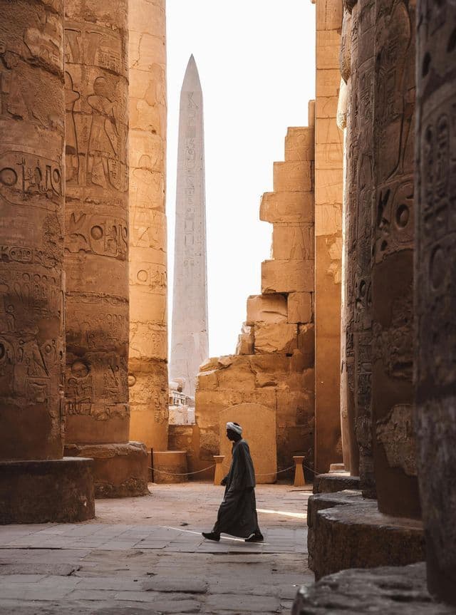 Un hombre con una túnica tradicional camina entre columnas de piedra masivas con jeroglíficos dentro de un templo, con un obelisco a lo lejos.
