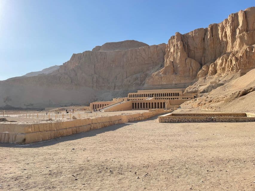 Un gran templo antiguo de múltiples terrazas, edificado en la base de un escarpado acantilado rocoso en un paisaje desértico bajo un cielo azul claro.