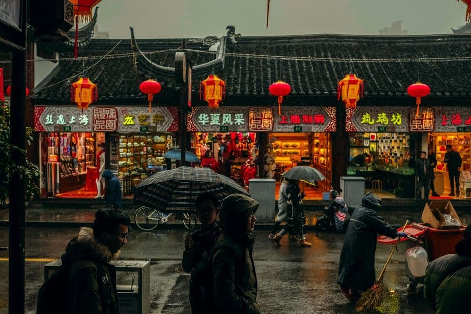 An einem regnerischen Tag gehen Menschen mit Regenschirmen auf einer nassen Straße, vorbei an hell erleuchteten Geschäften mit traditioneller Architektur und roten Laternen.
